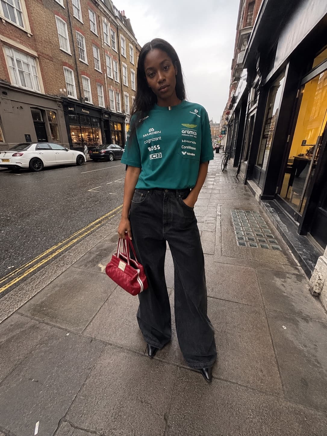 T-shirt del team Aston Martin di Formula 1 e jeans neri larghissimi, su un marciapiede bagnato a Londra.