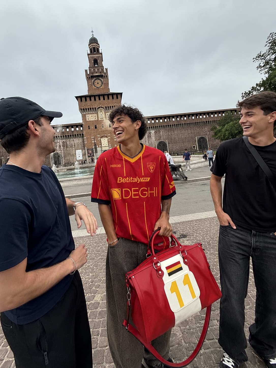 Tre amici ridono davanti al Castello Sforzesco: maglia del Lecce e borsone rosso numero 11.