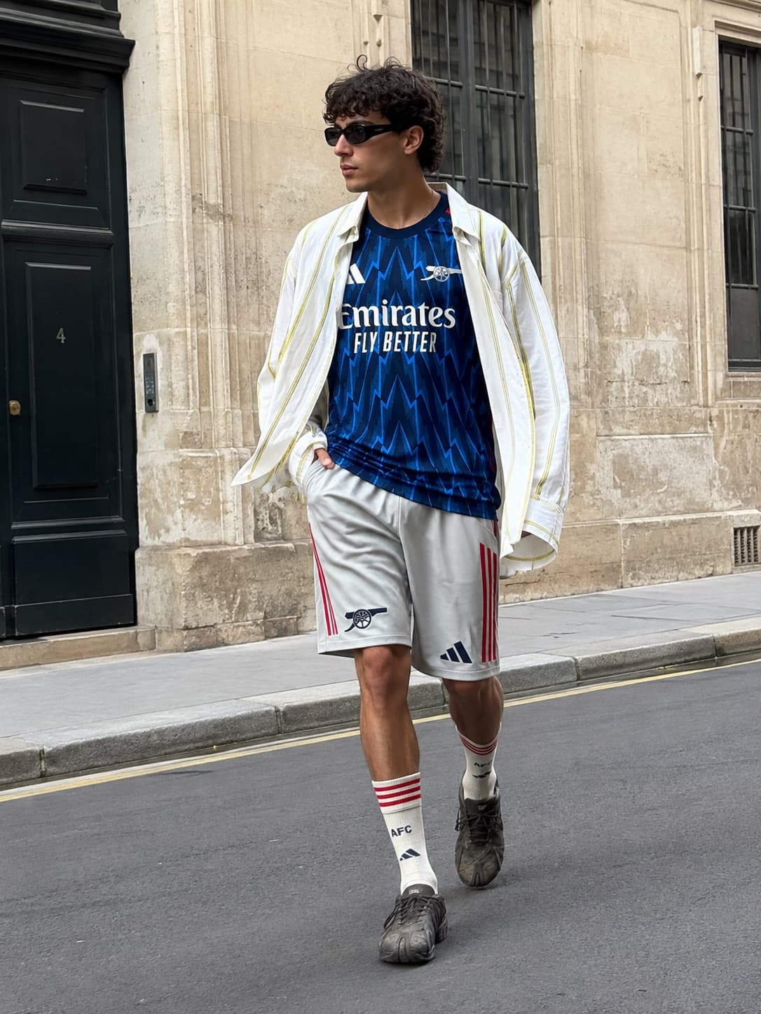 Maglia dell'Arsenal sotto una camicia bianca aperta, pantaloncini e calzettoni, mentre attraversa la strada.