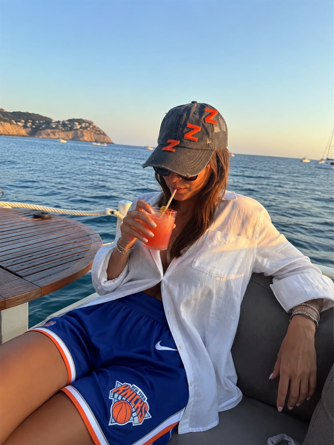 Pantaloncini dei Knicks e camicia di lino bianca, su una barca al tramonto con un drink in mano.